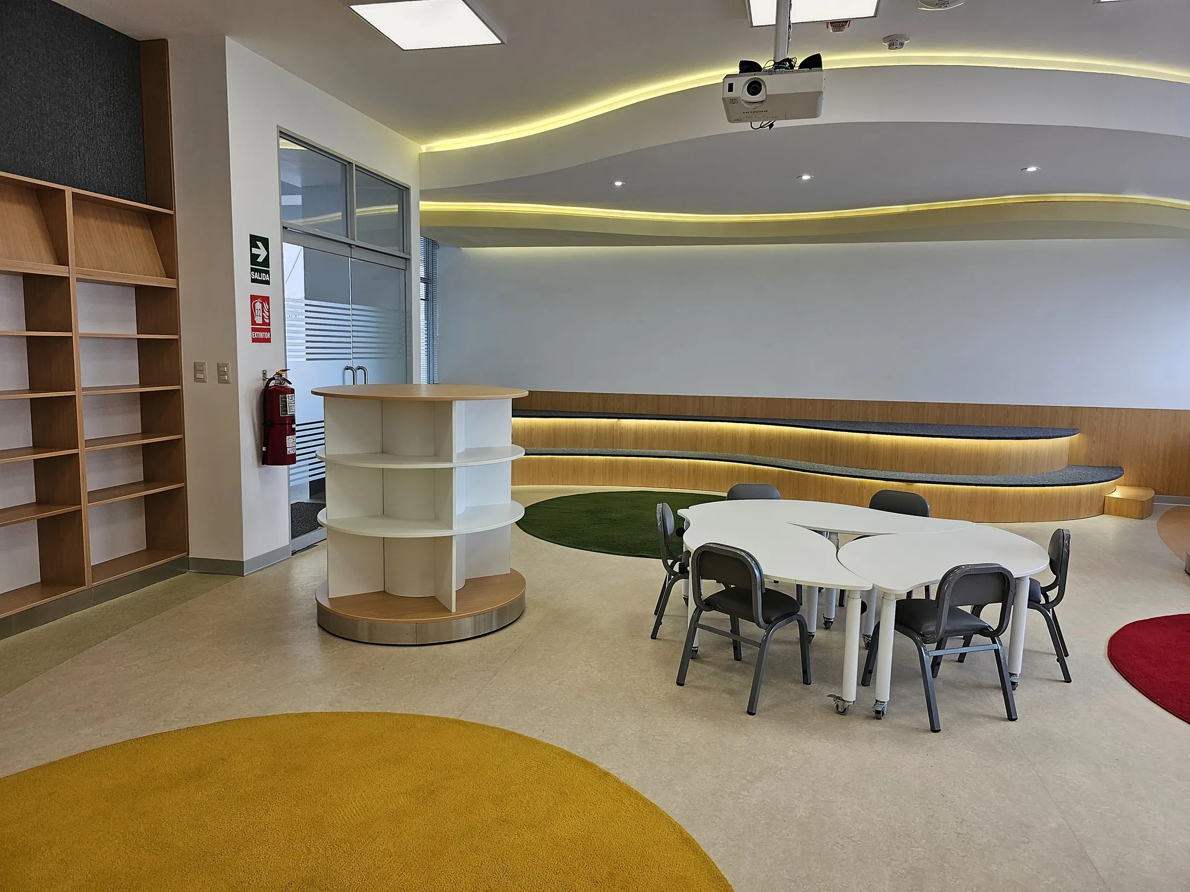 Biblioteca infantil con estantes de madera, un mueble circular, mesas blancas y alfombras redondas.
